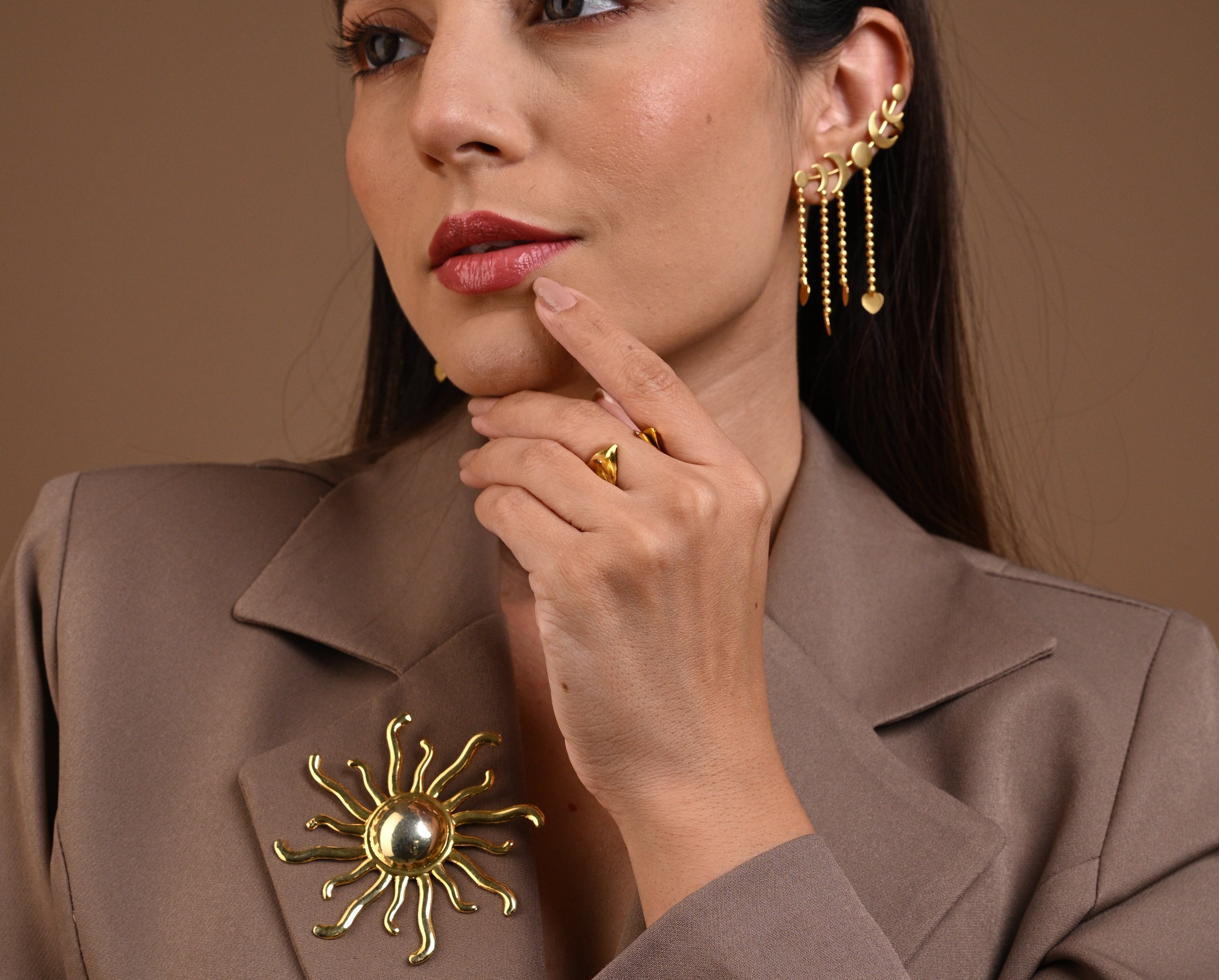 A close-up of a woman wearing gold jewelry, including a large sunburst brooch on her taupe blazer, a gold ring, and a long gold dangle earring, against a solid brown background.
